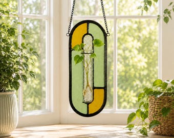 May include: A hanging stained glass plant propagation station featuring an oval black metal frame with yellow and light green glass panels. A clear glass test tube is centered within the frame, holding a small green plant cutting in water.