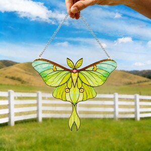 May include: A stained glass suncatcher shaped like a luna moth with translucent lime green and yellow wings, dark brown outlines, and a small pink center detail. The moth is suspended by a silver-toned metal chain for hanging display.