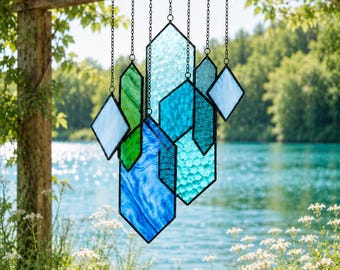 May include: A hanging stained glass mobile featuring multiple geometric shapes in shades of blue, green, and clear glass. The pieces are connected by thin dark metal chains, showcasing various textures including smooth, rippled, and honeycomb patterns.