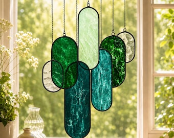 May include: A hanging stained glass suncatcher composed of multiple oval-shaped panels in varying shades of green, teal, and clear textured glass. Each panel is framed in black metal and suspended by thin chains at different vertical heights.