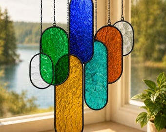 May include: A hanging suncatcher composed of seven vertical oval panels made of textured stained glass in blue, green, yellow, orange, teal, and clear tones. Each panel is framed in black metal and suspended by individual thin black chains.