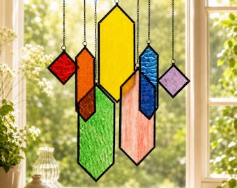 May include: A stained glass suncatcher featuring multiple geometric shapes in red, orange, yellow, green, blue, pink, and purple. Each piece is framed in black metal and suspended by thin black chains, creating a colorful hanging mobile display.