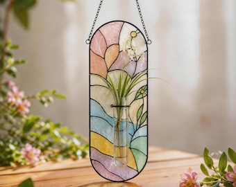 May include: A vertical oval wall hanging featuring a stained glass panel with pastel pink, blue, yellow, and green segments. A clear glass test tube is mounted in the center, holding a green air plant. The panel is suspended by a black metal chain.