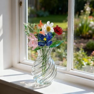 May include: A clear glass vase with a spiral design holds a colorful bouquet of glass flowers. The flowers include red, white, blue, orange, and pink blooms with green leaves. The vase sits on a white windowsill.
