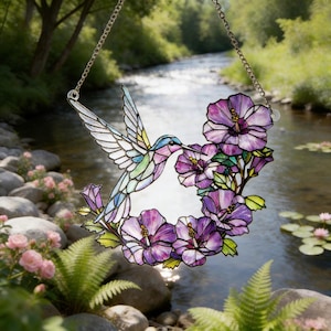 May include: Stained glass hummingbird and purple hibiscus flowers suncatcher hanging from a silver chain. The hummingbird has blue, green, and purple details. The flowers have purple petals and green leaves. The background shows a river and trees.
