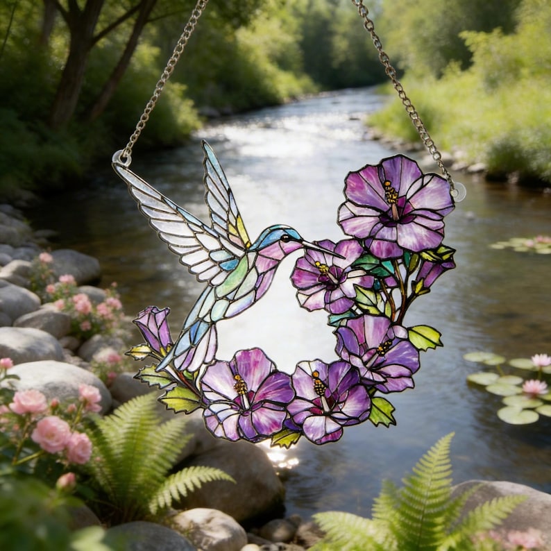 May include: Stained glass hummingbird and purple hibiscus flowers suncatcher hanging from a silver chain. The hummingbird has blue, green, and purple details. The flowers have purple petals and green leaves. The background shows a river and trees.