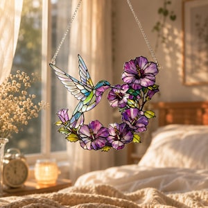 May include: A stained glass hummingbird with colorful wings and a long beak, hovering near a wreath of purple hibiscus flowers. The suncatcher is suspended by a silver chain, perfect for a window display.