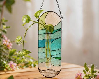 May include: A vertical oval stained glass wall hanging featuring a landscape of blue, green, and cream glass segments. A clear glass test tube is mounted in the center, holding a green pothos plant with trailing leaves and visible roots.