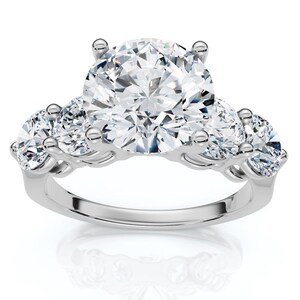 Puede incluir: Un anillo plateado con una gran piedra preciosa redonda y transparente en el centro, flanqueada por dos piedras preciosas más pequeñas y transparentes a cada lado. El anillo tiene una superficie pulida y reflectante.