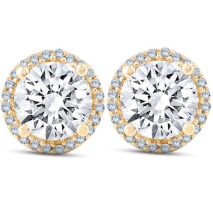 May include: A pair of round stud earrings featuring a large central clear stone surrounded by a halo of smaller clear stones. The stones are set in a yellow gold metal frame with four prominent prongs securing the center stone in place.
