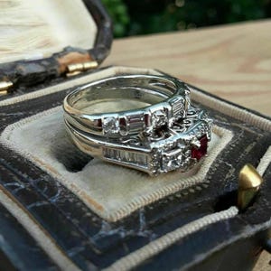 Peut inclure: Un ensemble de bagues de fiançailles en argent, avec une pierre centrale rouge et des diamants. L'ensemble est présenté dans un écrin ouvert. La bague présente un design détaillé et est accompagnée d'une alliance assortie.