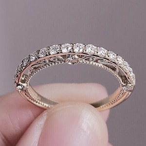 Könnte beinhalten: Ein goldfarbener Ring mit einem Band aus kleinen, funkelnden Edelsteinen. Der Ring weist aufwendige Filigranarbeiten an den Seiten und um die Edelsteine auf, wodurch eine Vintage-Ästhetik entsteht. Der Ring wird von einer Person gehalten.