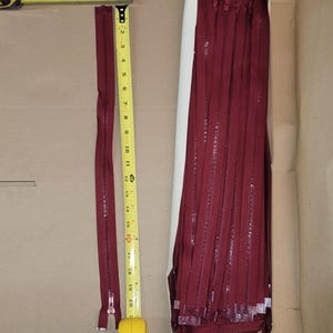 May include: A collection of burgundy zippers, some laid out next to a measuring tape. One zipper is fully extended, measuring approximately 20 inches. The zippers are neatly stacked in a cardboard box, ready for use in sewing or crafting projects.