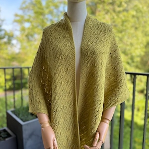 May include: A chartreuse knitted shawl featuring a textured pattern of diagonal lines and small raised bobbles. The garment is draped over a mannequin, showcasing an open front design with short sleeves and a consistent knit stitch throughout.