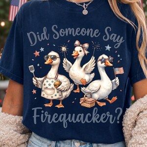 May include: Navy blue t-shirt with the text "Did Someone Say Firequacker?" and a graphic of three cartoon ducks celebrating with sparklers and an American flag. One duck wears sunglasses and an apron.