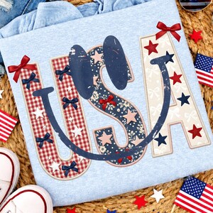 May include: Light blue USA graphic t-shirt with red, white, and blue accents. The letters are decorated with stars, bows, and floral patterns. The image includes white sneakers, denim shorts, sunglasses, and American flags.