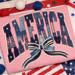 Könnte beinhalten: Ein rosa T-Shirt mit dem Wort "AMERICA" aus Denim-Patches, jeder Buchstabe mit weißen Sternen verziert. Eine Denim-Schleife mit rot-weißen Streifen akzentuiert das Design. Patriotisches Thema.