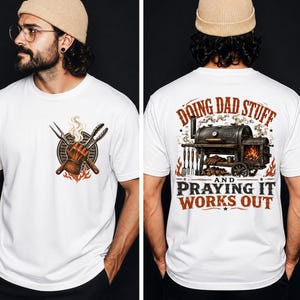 May include: A white t-shirt featuring a graphic on the front left chest of a gloved hand holding barbecue tongs and a fork over a grill. The back displays a large smoker grill with flames and the text DOING DAD STUFF AND PRAYING IT WORKS OUT.