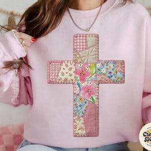 以下が含まれることがあります： パッチワークの十字架のデザインが施された、薄いピンク色のスウェットシャツ。十字架は、ピンク、白、青の色合いの様々な花柄と模様の生地で構成され、目に見える縫い目で縫い合わされています。スウェットシャツはクルーネックです。