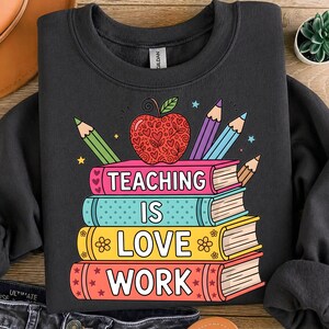 May include: Dark gray sweatshirt with a colorful graphic design. The design features a stack of books with the words "TEACHING IS LOVE WORK" and an apple, pencils, and stars. A great gift for teachers.