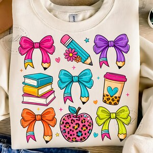 May include: Cream-colored sweatshirt featuring colorful school-themed graphics. Designs include bows, pencils, books, a coffee cup, and an apple with a leopard print pattern. The graphics are in bright pink, blue, orange, purple, and yellow.