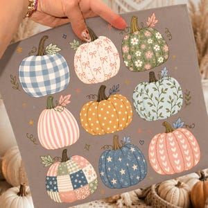 May include: A gray fabric square featuring a repeating pattern of nine pumpkins in various designs, including blue gingham, pink stripes, floral prints, polka dots, and heart motifs, accented with small stars and leaves throughout the illustration.