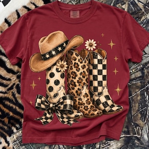 Puede incluir: Camiseta burdeos con un gráfico de un sombrero de vaquero y tres pares de botas de vaquero. Las botas tienen estampados de lunares, leopardo y cuadros. Estrellas doradas y acentos florales completan el diseño.