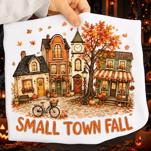 May include: A white fabric item featuring a colorful illustration of a small town street scene during autumn. The design includes buildings, a clock tower, a bicycle, pumpkins, and a large tree with orange leaves. The text reads SMALL TOWN FALL.