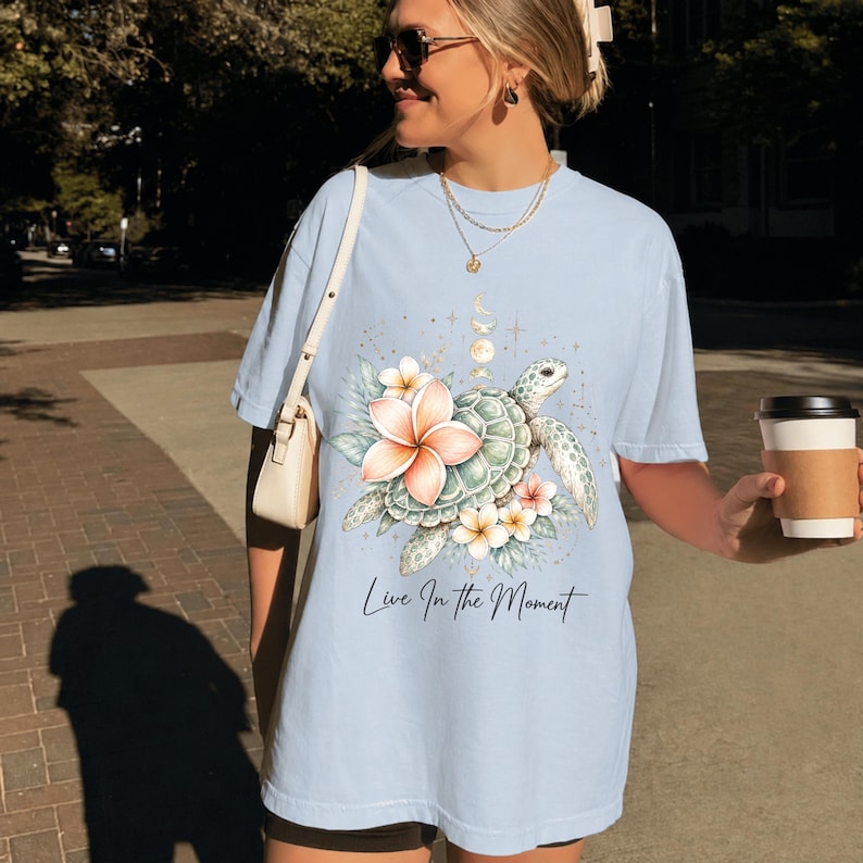 May include: A light blue short-sleeve t-shirt featuring a graphic print of a sea turtle surrounded by tropical plumeria flowers and celestial elements. The text "Live In The Moment" is printed in a black script font below the turtle illustration.