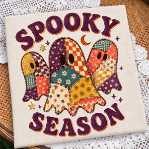 Puede incluir: Un artículo de tela de color blanco roto con forma cuadrada que presenta un estampado gráfico de tres fantasmas hechos de coloridos patrones de patchwork. El texto SPOOKY SEASON aparece en letras moradas con bordes naranjas y estrellas.