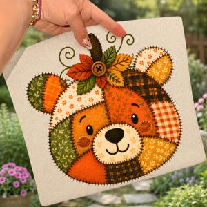 May include: A square decorative sign featuring a patchwork-style bear face illustration. The bear is composed of various orange, green, and brown patterned fabric textures with visible stitched outlines. A wooden button sits at the top center.