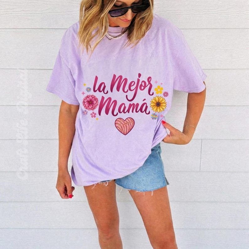 May include: A light purple t-shirt with the text "la Mejor Mam&aacute;" in pink script, decorated with floral and heart graphics. The shirt is a relaxed fit and is paired with denim shorts.