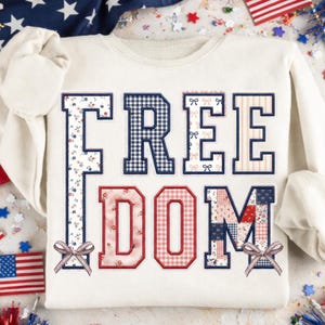 Może przedstawiać: Kremowa bluza z napisem "FREEDOM" w patchworkowym wzorze. Litery wykonane są z różnych wzorów, w tym kwiatowych, w kratkę i w paski, z małymi kokardkami. Odzież o tematyce patriotycznej.