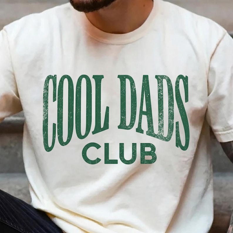May include: Cream-colored t-shirt with the text "COOL DADS CLUB" in distressed green lettering. The shirt has a crew neck and short sleeves. The design is centered on the chest.
