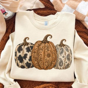 Pu&ograve; includere: Una felpa girocollo color crema con una stampa grafica di tre zucche. La zucca a sinistra presenta un motivo a macchie di mucca bianco e nero, quella centrale un disegno floreale marrone e quella a destra un motivo paisley nero.