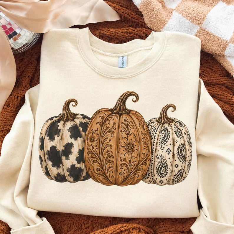 Pu&ograve; includere: Una felpa girocollo color crema con una stampa grafica di tre zucche. La zucca a sinistra presenta un motivo a macchie di mucca bianco e nero, quella centrale un disegno floreale marrone e quella a destra un motivo paisley nero.