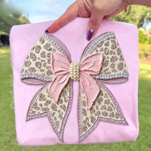 May include: A pink square object with a large, textured bow. The bow features a pink center, leopard print accents, and rhinestone trim. The bow is held by a hand with purple nail polish.