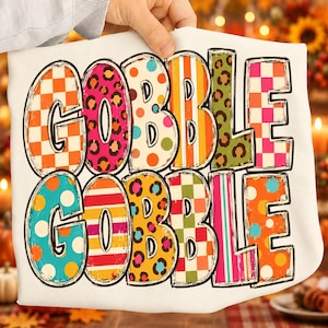 May include: A white t-shirt featuring the text GOBBLE GOBBLE in large, bold letters. Each letter is filled with a different colorful pattern, including orange and white checkers, pink leopard print, orange polka dots, stripes, and green leopard print.