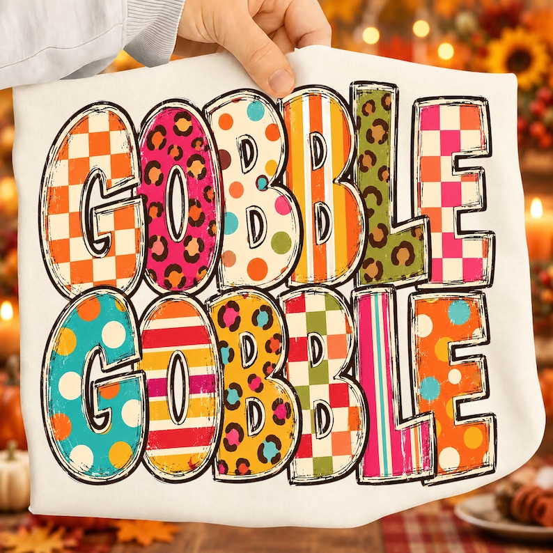 May include: A white t-shirt featuring the text GOBBLE GOBBLE in large, bold letters. Each letter is filled with a different colorful pattern, including orange and white checkers, pink leopard print, orange polka dots, stripes, and green leopard print.