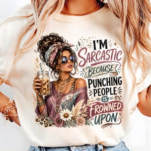 May include: A cream-colored t-shirt featuring a graphic of a person with a messy bun, sunglasses, and layered necklaces holding an iced coffee. The text reads, I'M SARCASTIC BECAUSE PUNCHING PEOPLE IS FROWNED UPON, surrounded by floral and leopard accents.