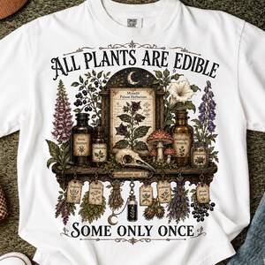 May include: A white t-shirt featuring a graphic print of a botanical display with the text ALL PLANTS ARE EDIBLE SOME ONLY ONCE. The design includes apothecary bottles, a skull, mushrooms, and hanging herb labels with plant names.