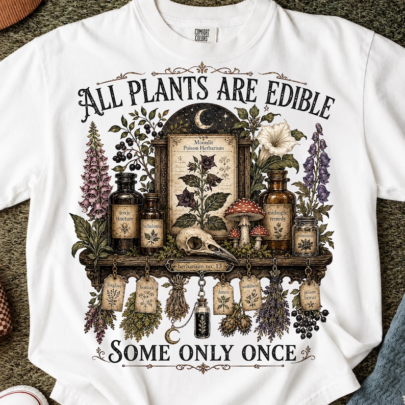 May include: A white t-shirt featuring a graphic print of a botanical display with the text ALL PLANTS ARE EDIBLE SOME ONLY ONCE. The design includes apothecary bottles, a skull, mushrooms, and hanging herb labels with plant names.