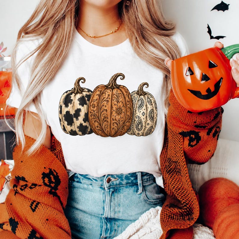 Pu&ograve; includere: Una t-shirt bianca a maniche corte con una stampa grafica di tre zucche. La zucca a sinistra ha una stampa maculata bianca e nera, quella centrale un complesso motivo floreale marrone e quella a destra un design paisley color crema e nero.