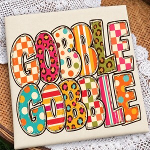 May include: A square white sign featuring the text GOBBLE GOBBLE in large, bold letters. Each letter is filled with a different colorful pattern, including orange and white checkers, pink leopard print, polka dots, stripes, and green leopard print.