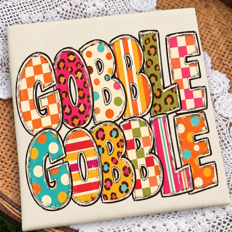 May include: A square white sign featuring the text GOBBLE GOBBLE in large, bold letters. Each letter is filled with a different colorful pattern, including orange and white checkers, pink leopard print, polka dots, stripes, and green leopard print.