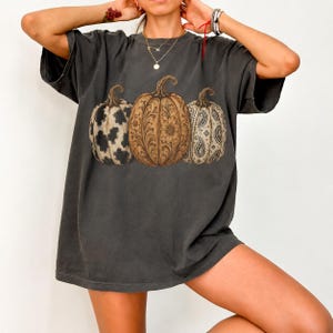 Pu&ograve; includere: Una t-shirt a maniche corte grigio antracite con una stampa grafica di tre zucche. La zucca a sinistra ha una stampa muccata bianca e nera, quella centrale un motivo floreale marrone e quella a destra un design paisley bianco e nero.