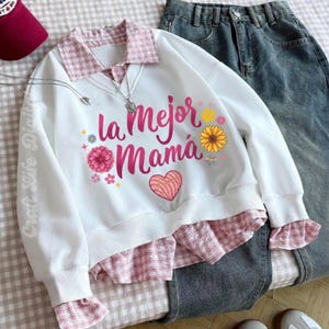 May include: White sweatshirt with the text "la Mejor Mam&aacute;" in pink, floral accents, and a heart design. The sweatshirt has a pink plaid collar and cuffs, paired with denim jeans. A maroon baseball cap and necklace are also visible.
