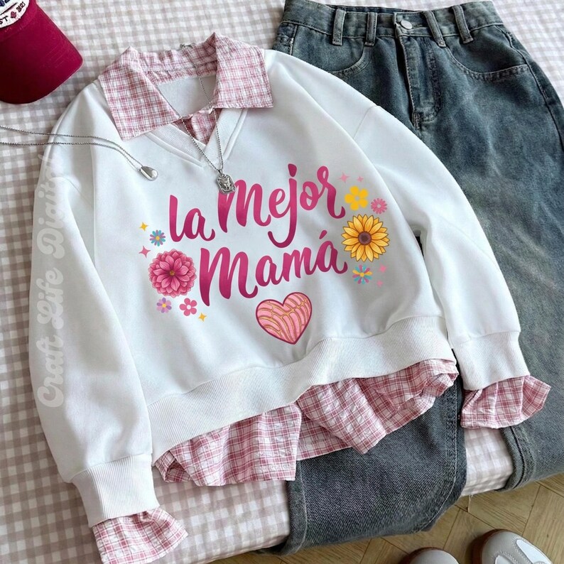 May include: White sweatshirt with the text "la Mejor Mam&aacute;" in pink, floral accents, and a heart design. The sweatshirt has a pink plaid collar and cuffs, paired with denim jeans. A maroon baseball cap and necklace are also visible.