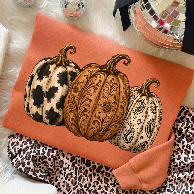 Pu&ograve; includere: Una felpa arancione brillante con una grafica raffigurante tre zucche. La zucca a sinistra ha una stampa muccata bianca e nera, quella centrale un motivo floreale marrone e quella a destra una stampa paisley bianca e nera.