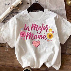 May include: White t-shirt with the text "la Mejor Mam&aacute;" in pink script, decorated with a pink flower, a sunflower, and a heart-shaped concha. The shirt is displayed on a wooden surface.